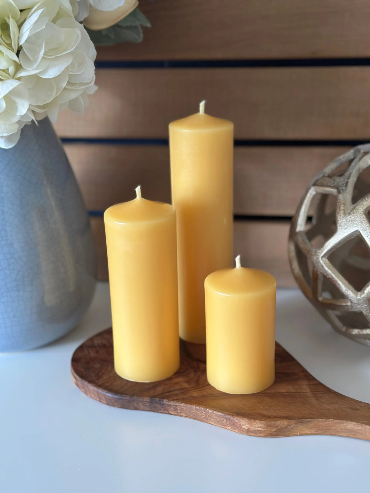 Three narrow candles on a wooden tray with a vase of flowers and decorative object in the background.